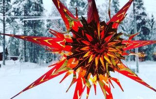 En levande, stjärnformad prydnad med röda och guldiga spikar hänger mot en snöig utomhusbakgrund, med snötäckta träd och ett vintrigt landskap. Prydnaden lyser varmt, i kontrast till det kalla landskapet.