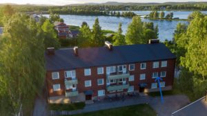 Flygfoto över en trevånings tegelbyggnad med balkonger, omgiven av träd. Cyklar är parkerade utanför. Det finns en älv och fler hus i bakgrunden under en blå himmel med moln.