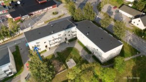 Flygfoto över ett litet bostadsområde med två intilliggande L-formade hyreshus omgivna av träd. En lekplats syns på den gemensamma gården mellan byggnaderna. Vägar och fler hus syns i närheten.