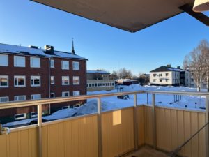 Utsikt från en balkong som visar en snöig scen med tegel och vita byggnader under en klarblå himmel. Området är lugnt och några kala träd ligger utspridda. Balkongen har gult räcke och heltäckningsmatta.