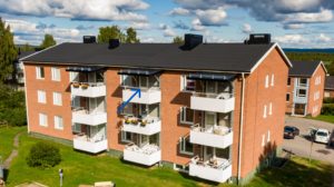 Ett hyreshus i tegel med tre våningar, med vita balkonger. En blå pil pekar mot ena balkongen på andra våningen, som har blomkrukor och möbler. Byggnaden är omgiven av en gräsyta och andra liknande strukturer.