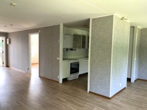 En lägenhet med öppenplanlösning, med grå tapeter på väggarna samt ett träfärgat golv. Rakt fram skymtar köksdelen med vita skåpdörrar, en spis och spisfläkt. en öppnad innerdörr leder till ett sovrum där solljuset strilar in.