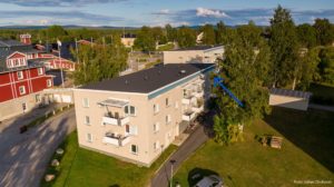 Flygfoto över ett litet bostadsområde med två intilliggande ljusa hyreshus omgivna av träd och grönområden. Vägar och fler hus syns i närheten. En blå pil pekar mot en specifik lägenhet i det främre hyreshuset