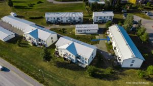 Flygfoto över ett bostadsområde med flera vita byggnader omgivna av grönt gräs och träd. En blå pil pekar mot en av byggnaderna.