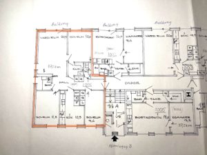 Handritad planlösning för ett flerfamiljshus med flera markerade rum och mått. En central lägenhet är markerad med orange markering, vilket markerar dess rum och planlösning. Handskrivna anteckningar och siffror syns.