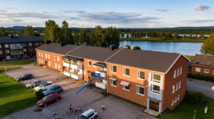 Ett tvåvåningshus i tegel med vita balkonger. En pil pekar mot en balkong på andra våningen. Byggnaden är omgiven av en asfalterad parkering, träd och en älv, samt en delvis molnig himmel.