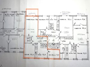 Handritad planlösning för ett flerfamiljshus med flera markerade rum och mått. En central lägenhet är markerad med orange markering, vilket markerar dess rum och planlösning. Handskrivna anteckningar och siffror syns.