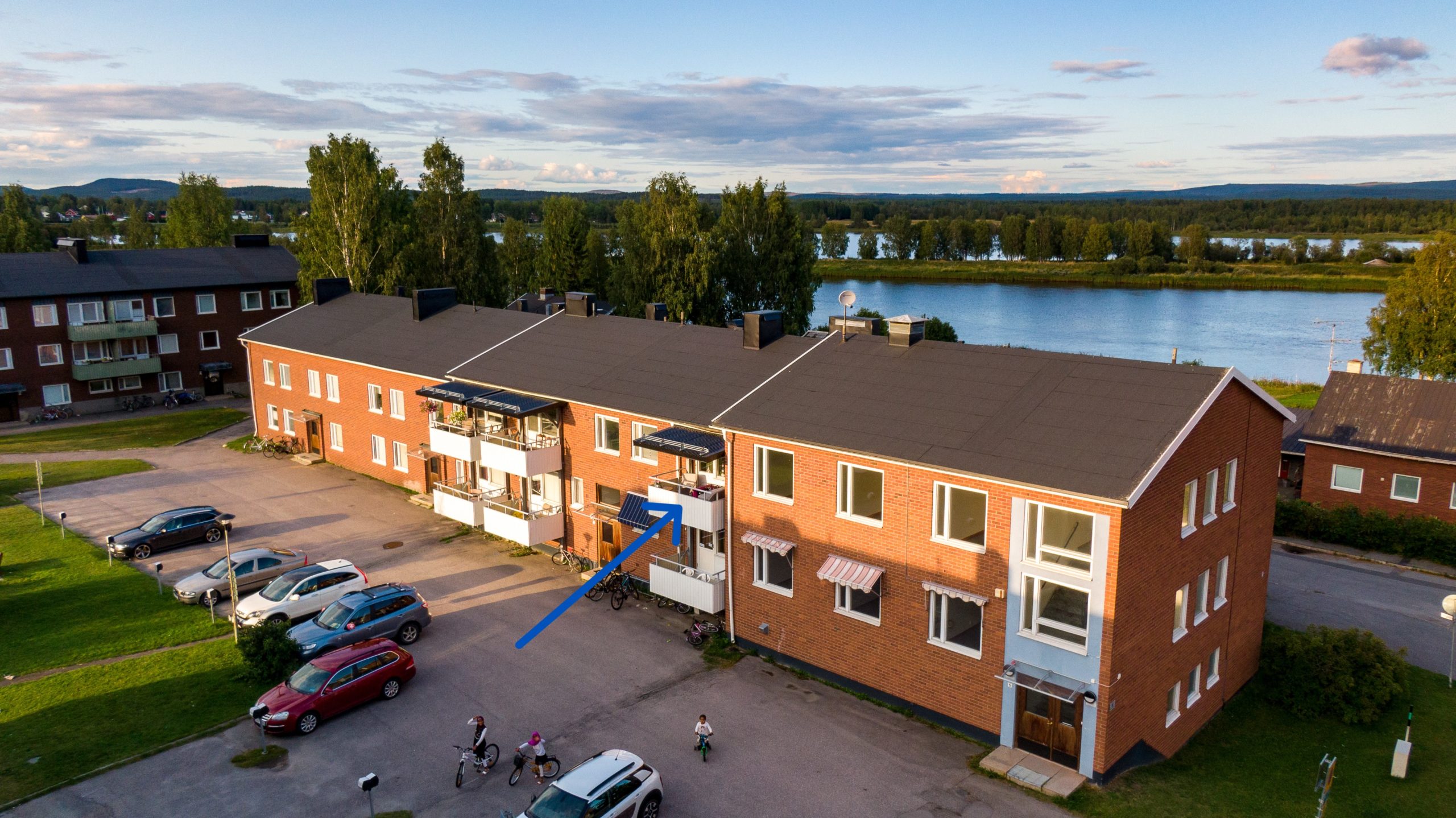 Ett tvåvåningshus i tegel med vita balkonger. En pil pekar mot en balkong på andra våningen. Byggnaden är omgiven av en asfalterad parkering, träd och en älv, samt en delvis molnig himmel.