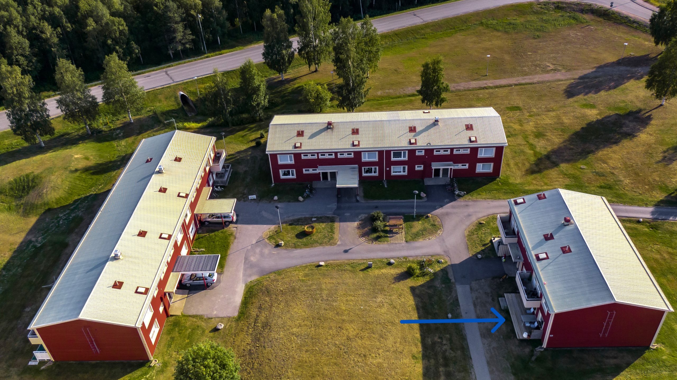 Flygfoto över tre röda byggnader nära en väg, omgiven av träd och gräsytor. En pil markerar en särskild lägenhet i en av fastigheterna.