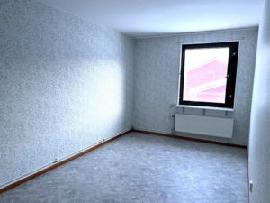 Sovrum med blommiga tapeter och gråfärgat golv. Ett fönster på den bakre väggen släpper in ljus.