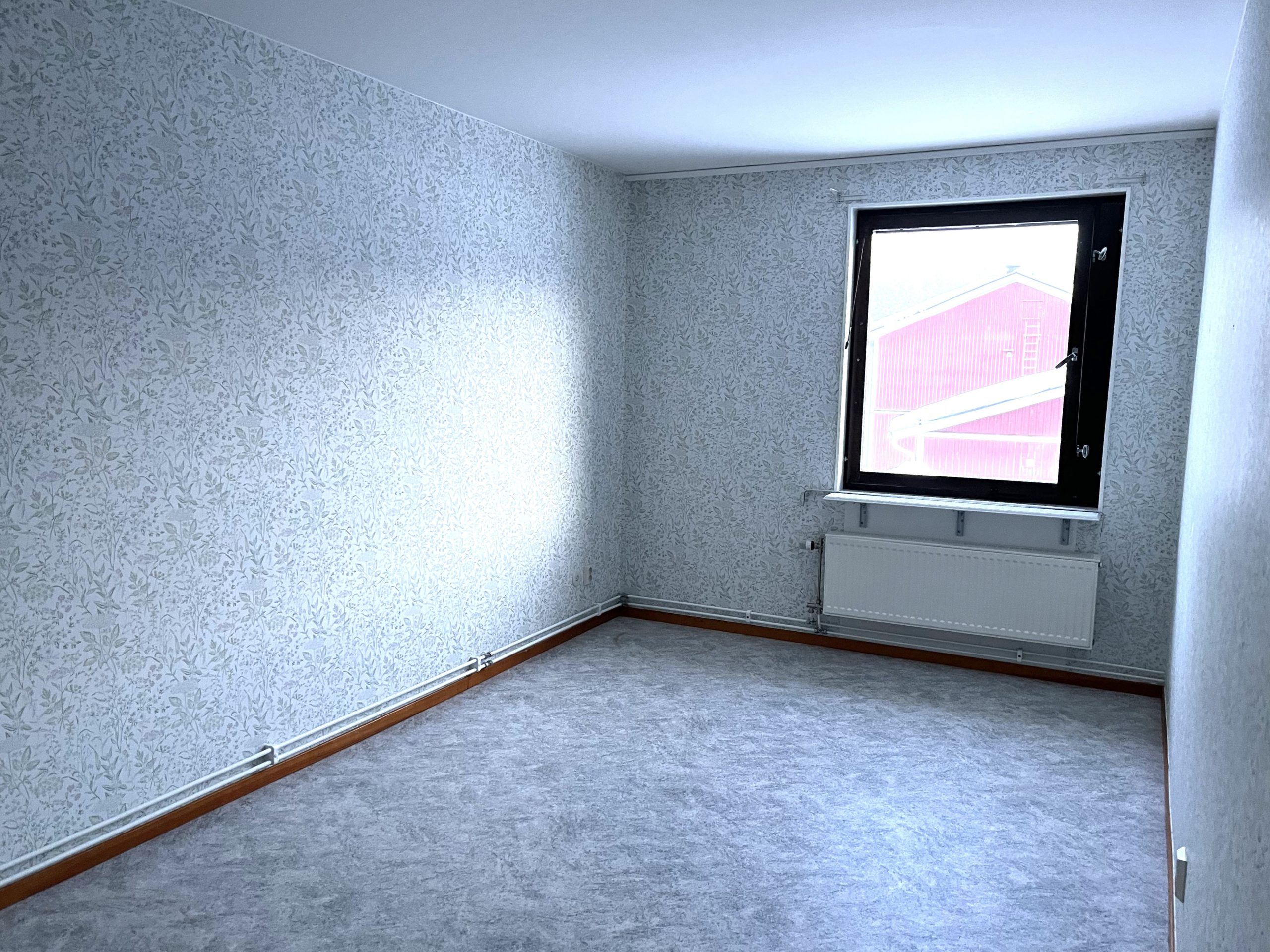 Sovrum med blommiga tapeter och gråfärgat golv. Ett fönster på den bakre väggen släpper in ljus.