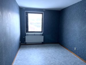 Sovrum med blå tapeter och gråfärgat golv. På det bakre väggen finns ett fönster som släpper in ljus.