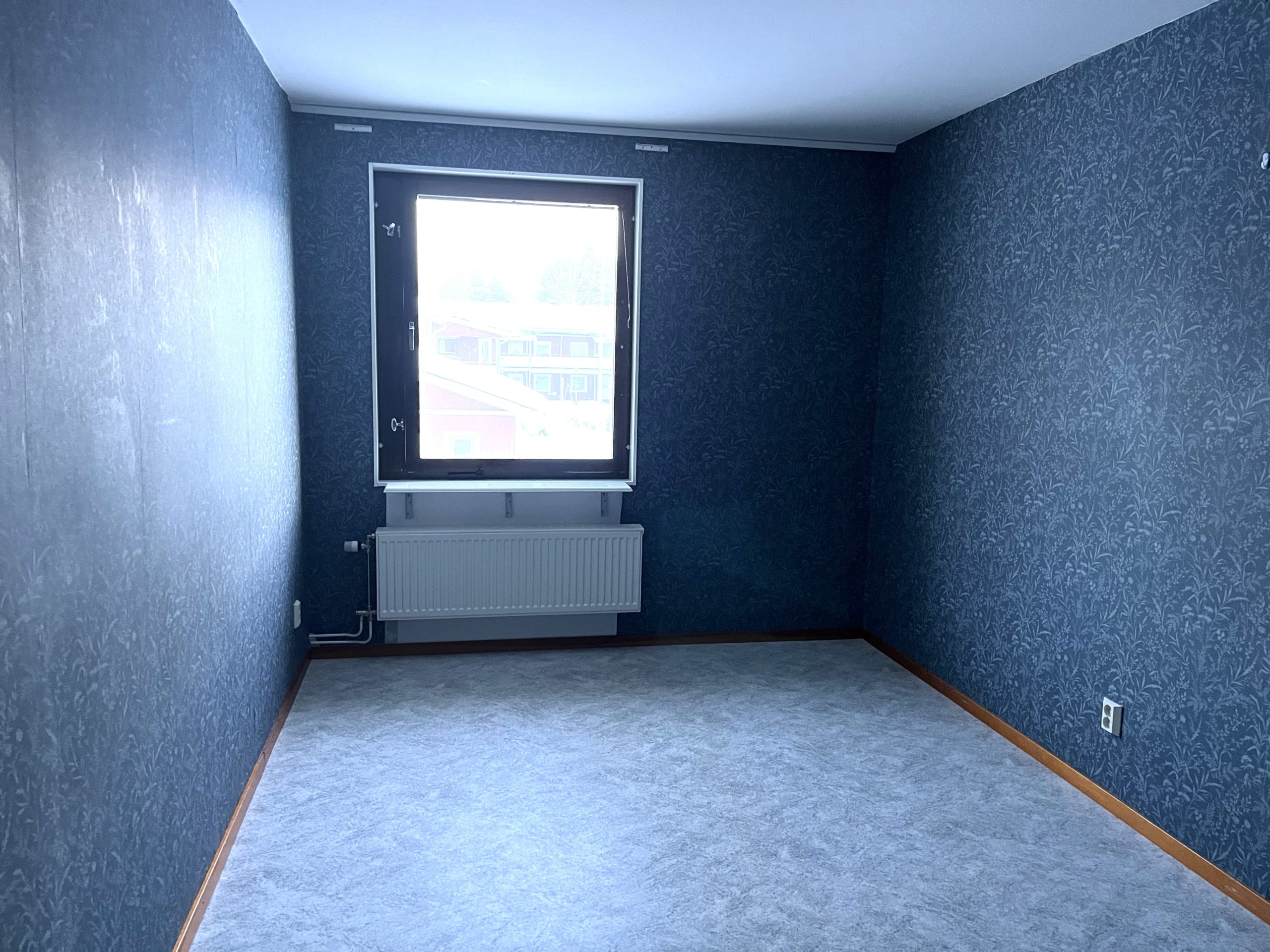Sovrum med blå tapeter och gråfärgat golv. På det bakre väggen finns ett fönster som släpper in ljus.