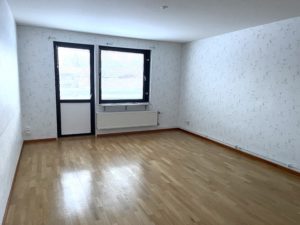 Vardagsrum med ljusa, blommiga tapeter och ett parettgolv. Ett fönster och balkongdörr på den bakre väggen släpper in ljus.