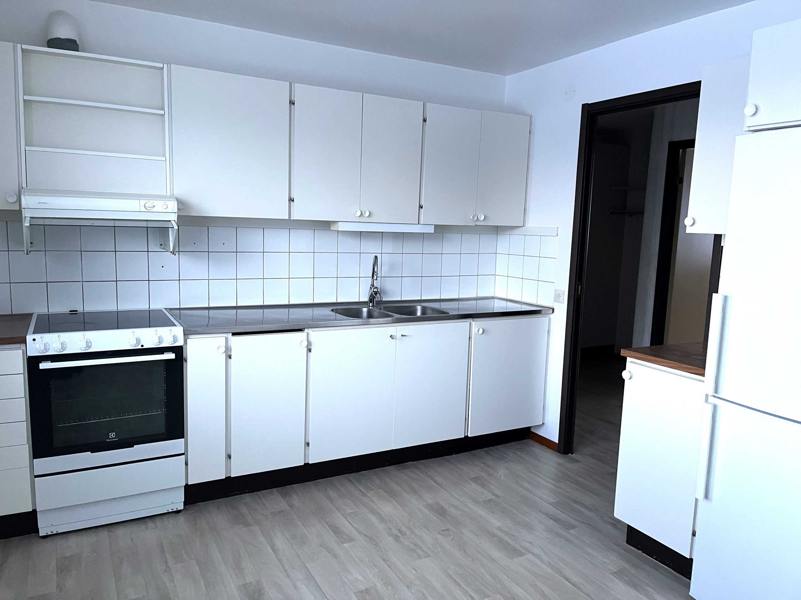 Kök med vita skåpluckor, vitt kakel samt ett träfärgat golv. En dörröppning leder ut till lägenhetens hall.