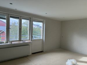 Ett vardagsrum med ljusa väggar, tre fönster och en balkongdörr släpper in dagsljus. Renoveringsarbete pågår.