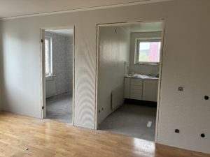 Bilden visar ett vardagrum i förgrunden med parkettgolv och ljusa väggar. Två dörröppningar leder till ett sovrum och ett kök som genomgår renovering.