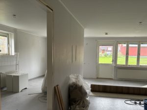 Lägenhet med öppen planlösning där renoveringsarbete pågpr. Ljusa väggar, träfärgat golv, tre fönster samt en balkongdörr i bakgrunden släpper in dagsljus.