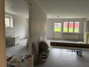 Lägenhet med öppen planlösning där renoveringsarbete pågpr. Ljusa väggar, träfärgat golv, tre fönster samt en balkongdörr i bakgrunden släpper in dagsljus.