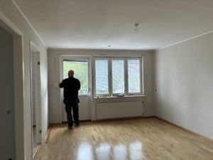 Vardagsrum med parkettgolv och ljusa väggar. Längst bort i bild tre fönster och en balkongdörr som släpper in dagsljus. Framför balkongdörren står en svartklädd person som fixar med persiennerna.