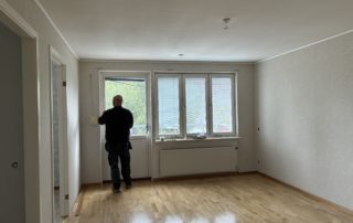 Vardagsrum med parkettgolv och ljusa väggar. Längst bort i bild tre fönster och en balkongdörr som släpper in dagsljus. Framför balkongdörren står en svartklädd person som fixar med persiennerna.