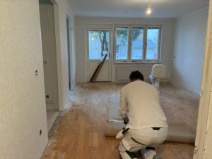 Renoveringsarbete i en lägenhet. I förgrunden en hantverkare i ljusa kläder som rullar ihop täckpapper.