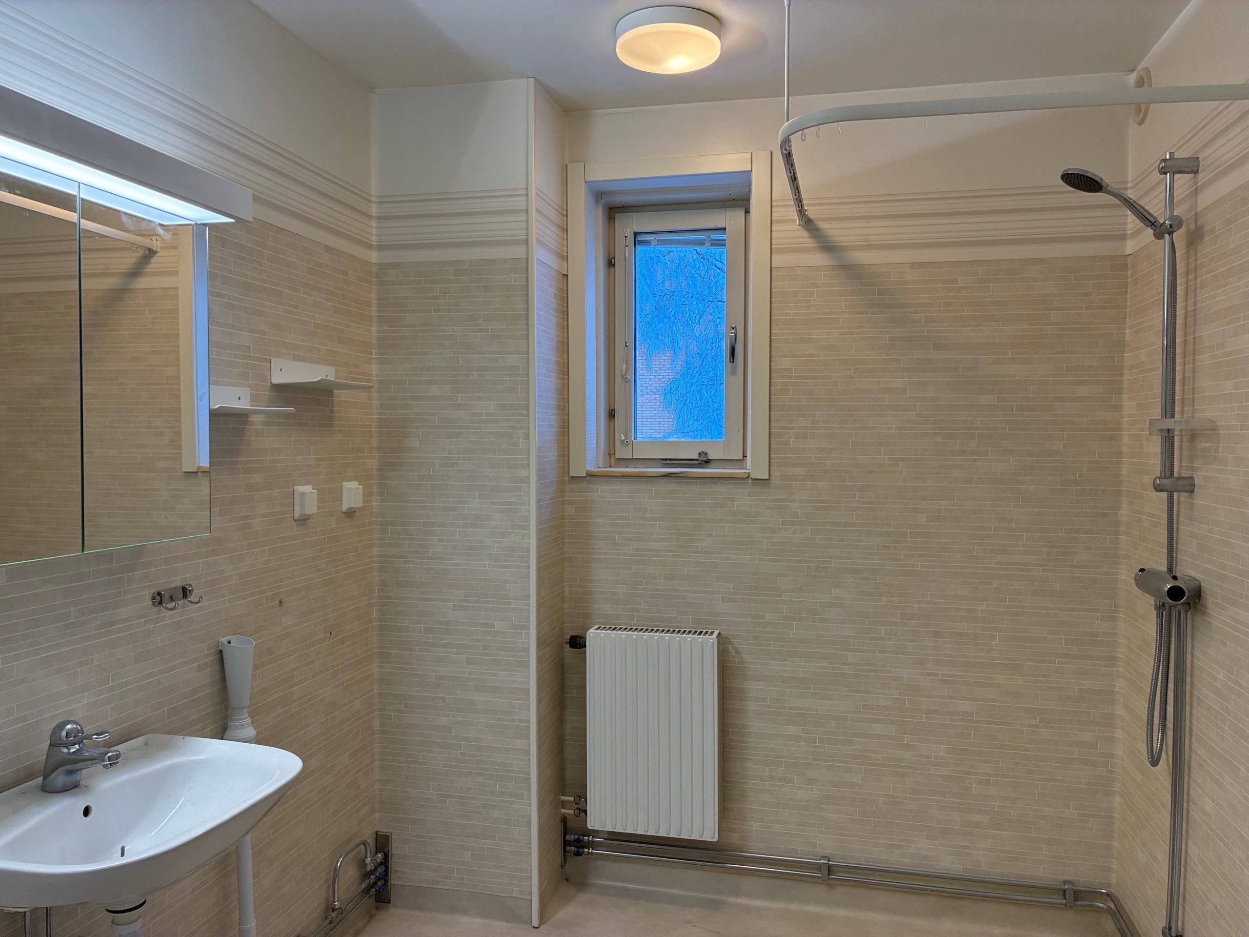 Badrum i beiga toner med duschhörna. Ett långsmalt fönster släpper in dagsljus. Ett tvättställ och badrumsskåp skymtar till vänster i bild.