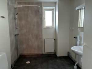 Badrum med ljusgrå duschhörna, vita väggar samt en mörkgrå golvmatta. På den bare väggen finns ett långsmalt fönster som släpper in ljus.