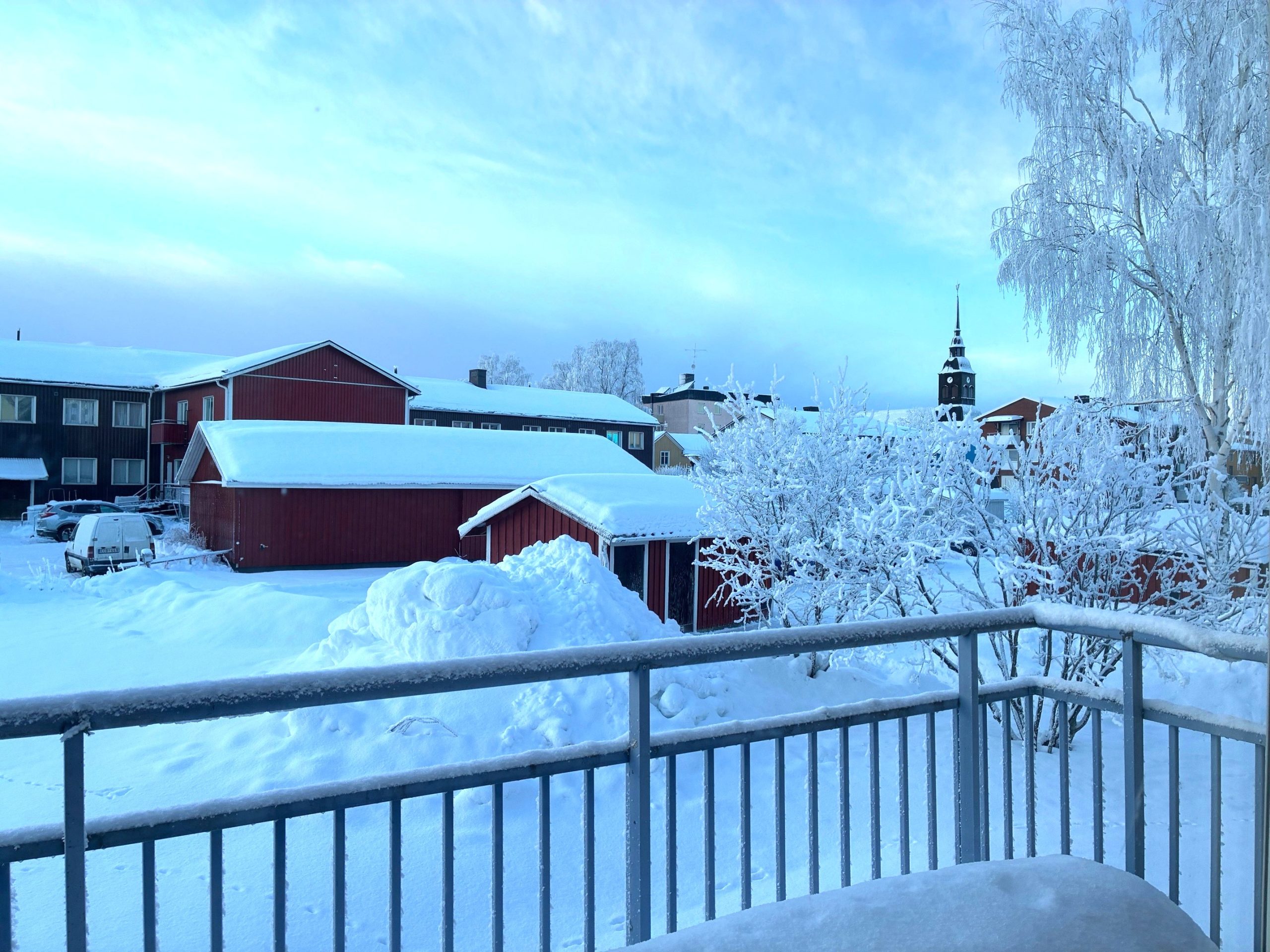 Utsikt från en balkong med snö, byggnader och ett kyrktorn.