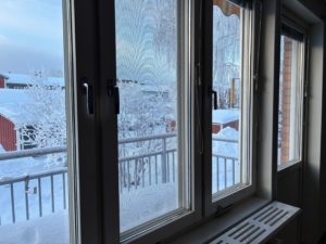 Utsikt från fönster med en balkong i förgrunden. Utomhus skymtar byggnader i ett snötäckt landskap.