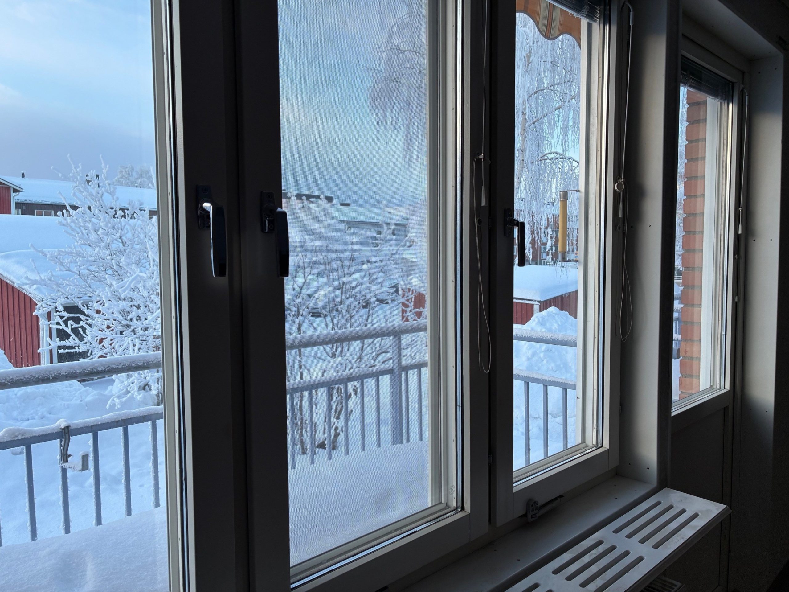 Utsikt från fönster med en balkong i förgrunden. Utomhus skymtar byggnader i ett snötäckt landskap.
