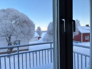 Utsikt från fönster med en balkong i förgrunden. Utom hus skymtar ett snötäckt landskap med ett hus och träd.