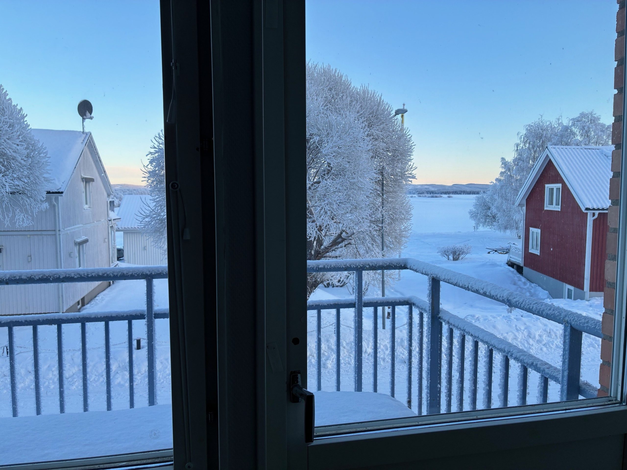 Utsikt från ett fönster med en balkong i förgrunden. Det skymtar två villor samt träd och en älv.