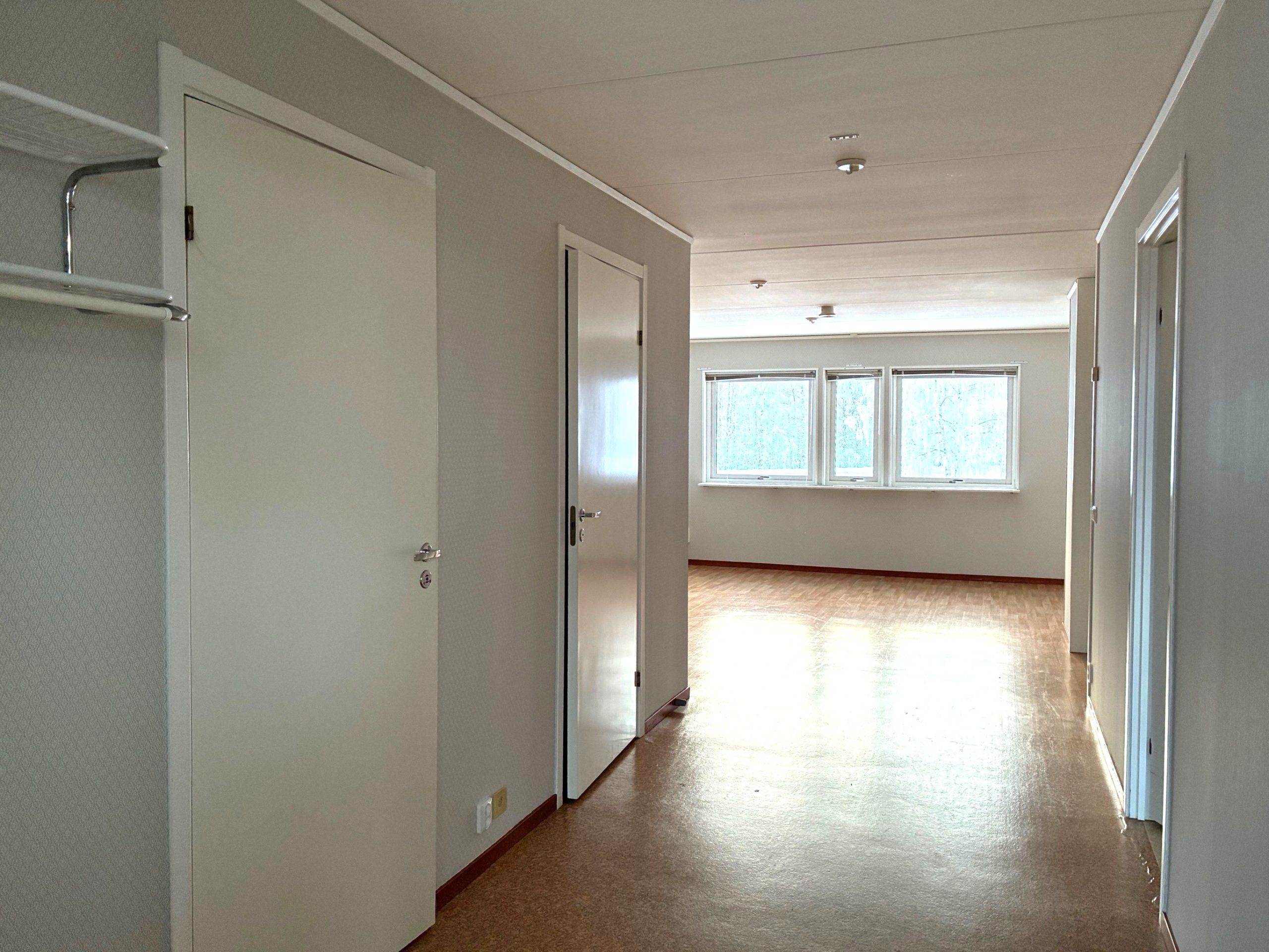 Hall och vardagsrum i en lägenhet med öppen planlösning. Ljusa väggar och ett brunt golv. På den bakre väggen finns fönster som släpper in dagsljus.