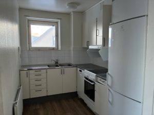 Ett kompakt kök med ljus köksinredning, vita väggar samt ett träfärgat golv. Ett mindre fönster sitter ovanför diskbänken och släpper in ljus.