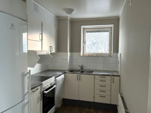 Ett kompakt kök med vit köksinredning, vita väggar samt ett träfärgat golv. Ovanför diskbänken finns ett mindre fönster som släpper in dagsljus,