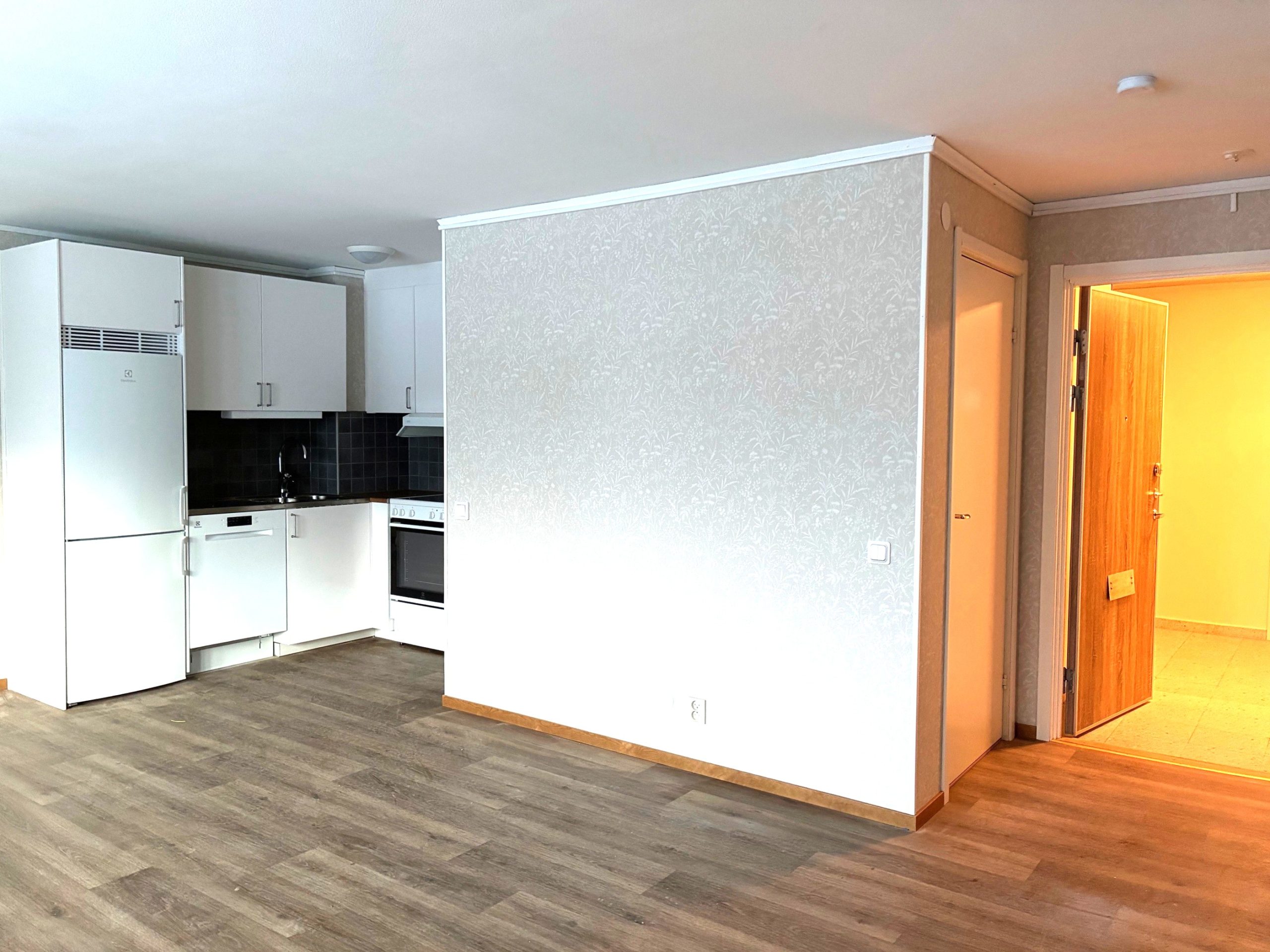 Delar av ett kök, allrum och hall i en mindre lägenhet med öppen planlösning. Ljusa väggar och ett träfärgat golv. Köksinredningen är vit.