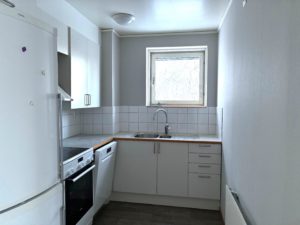 Ett kompakt kök med vit köksinredning och ljusa väggar. Ett fönster ovanför diskbänken släpper in dagsljus.