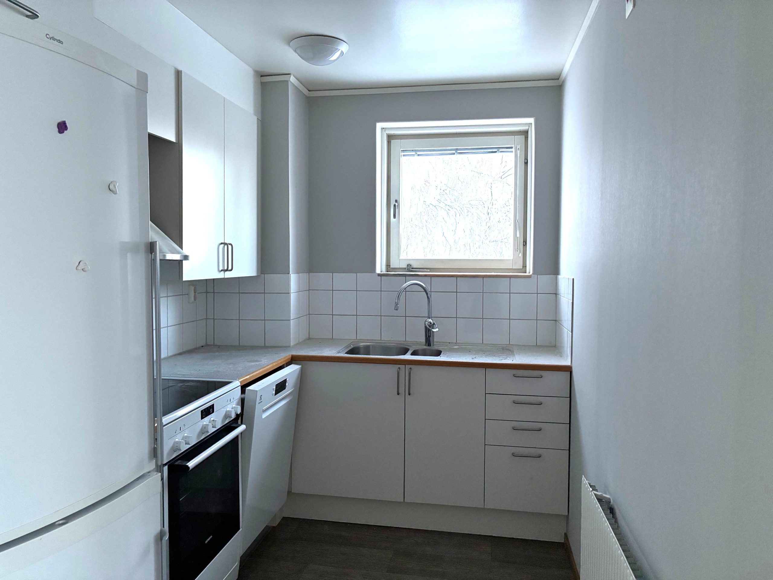 Ett kompakt kök med vit köksinredning och ljusa väggar. Ett fönster ovanför diskbänken släpper in dagsljus.