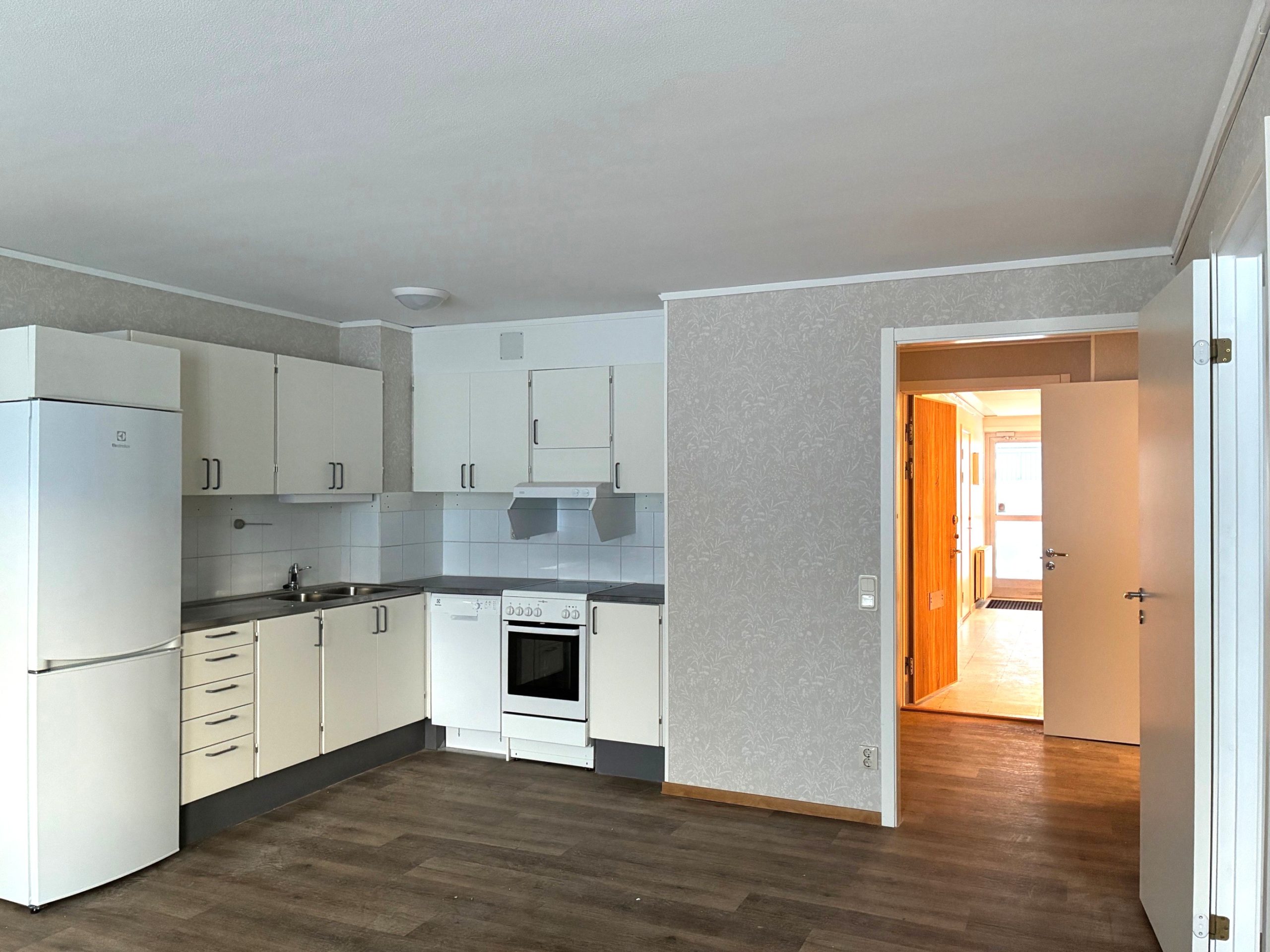Del av kök, samt hall av en mindre lägenhet med öppen planlösning. Vit köksinredning, ljusa väggar, samt träfärgat golv. i bakgrunden syns lägenhetsdörren och trapphuset.