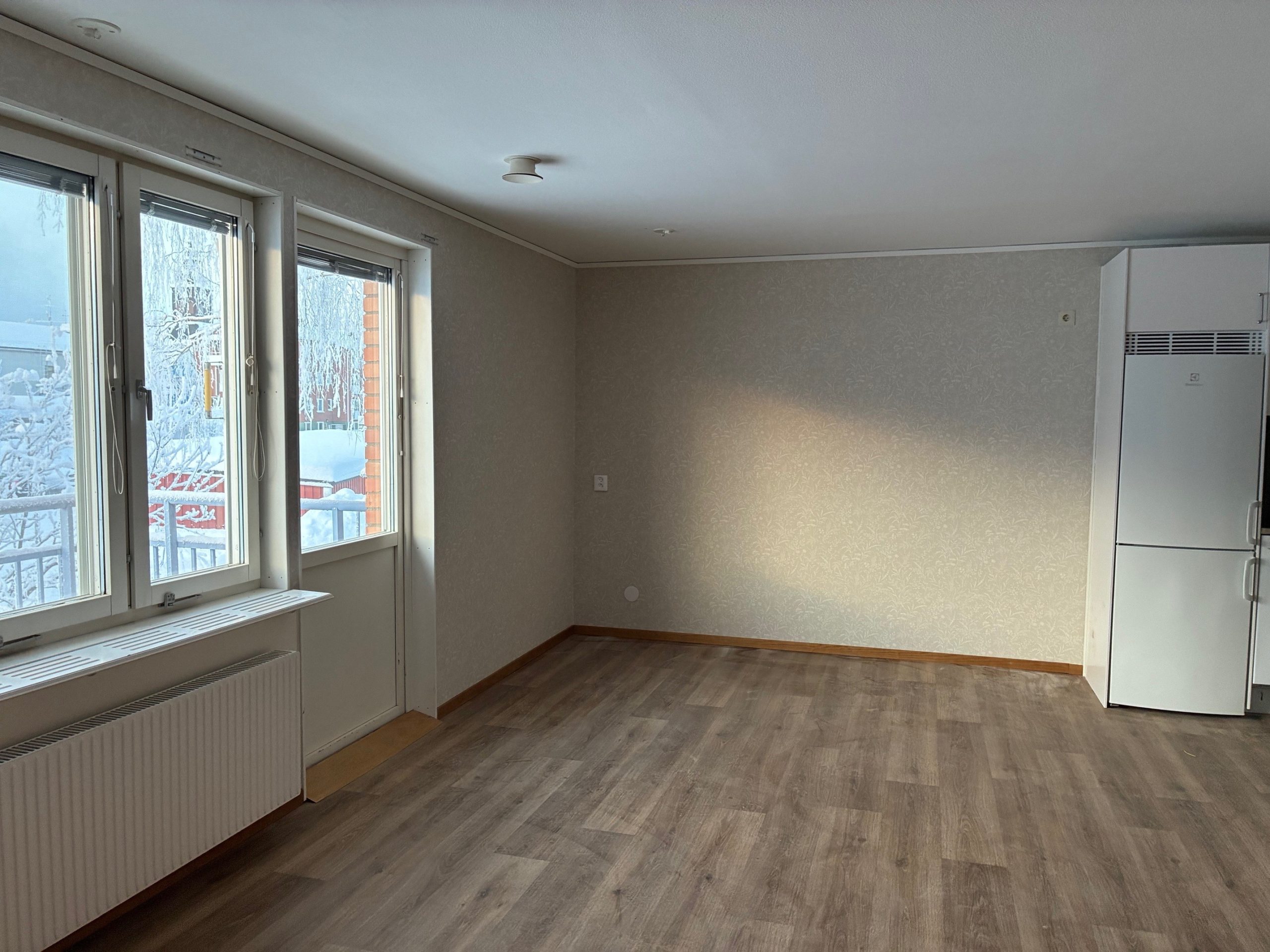 Del av allrummet i en mindre lägenhet med öppen planlösning. Ljusa väggar och ett träfärgat golv. Till vänster i bild skymtar fönster och en balkongdörr.