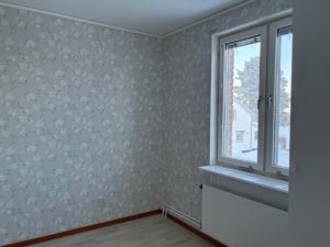 Ett mindre sovrum med blommiga tapeter och träfärgat golv. Ett fönster finns till höger i bild.