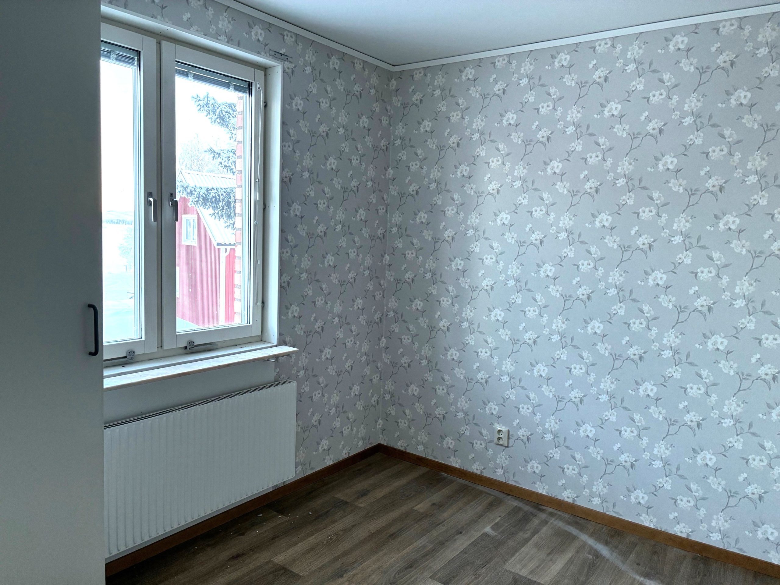 Ett mindre sovrum med blommiga tapeter och träfärgat golv. Ett fönster finns till vänster i bild.