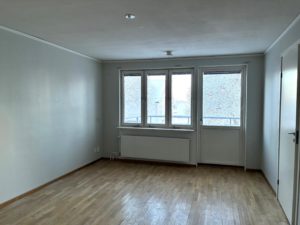 vardagsrum med ljusa tapeter och parkettgolv. Fönster och en balkongdörr på den bakre väggen släpper in ljus.