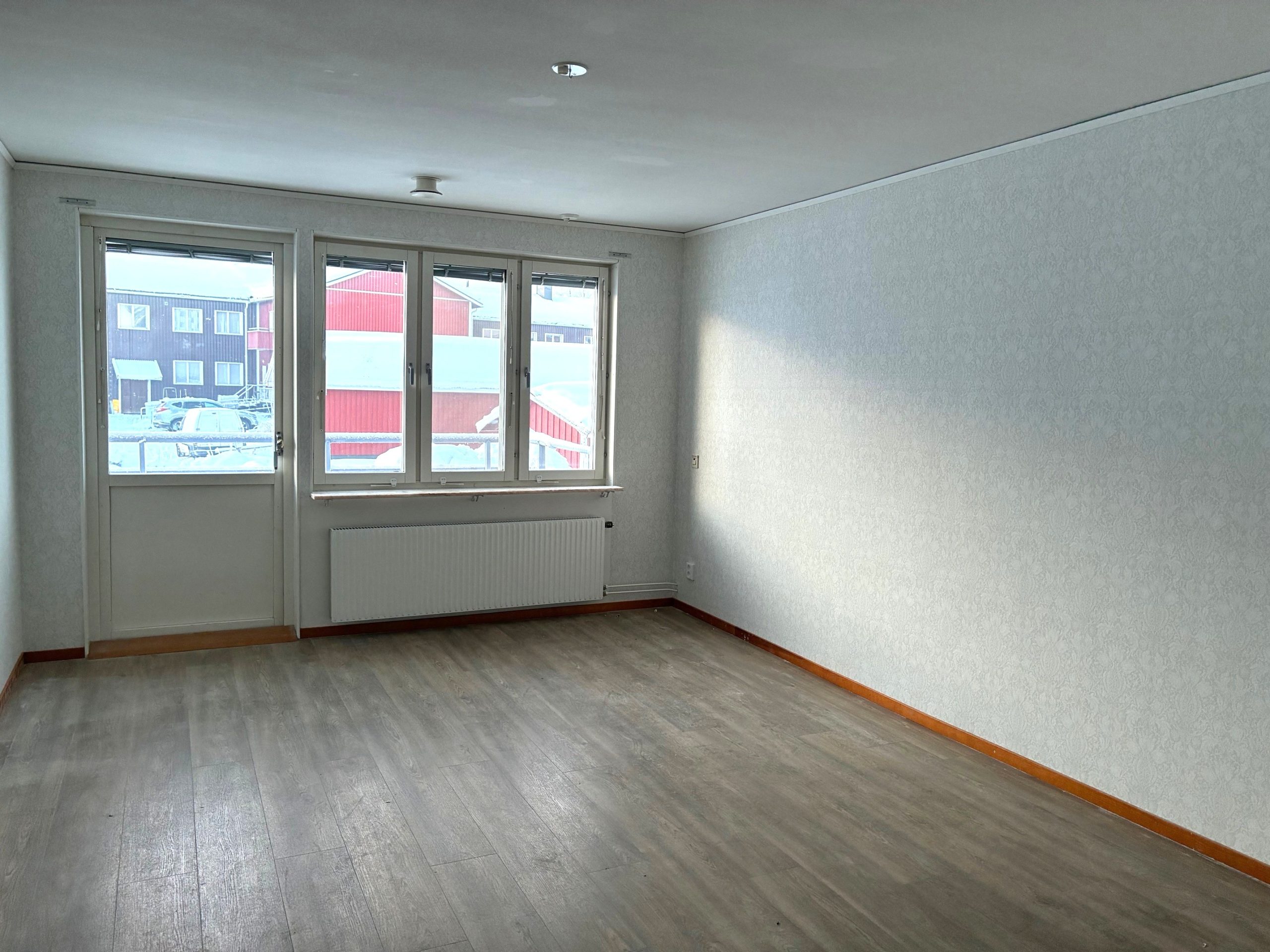 Vardagsrum med ljusa väggar och ett träfärgat golv. På den bakre väggen finns fönster och en balkongdörr som släpper in dagsljus.