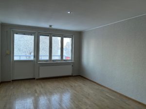 Ett vardagsrum med ljusa tapeter och parkettgolv. Fönster och en balkongdörr syns på den bakre väggen och släpper in dagsljus.