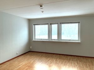 vardagsrum med ljusa tapeter och ett brunt golv. Flera fönster släpper in dagsljuset.