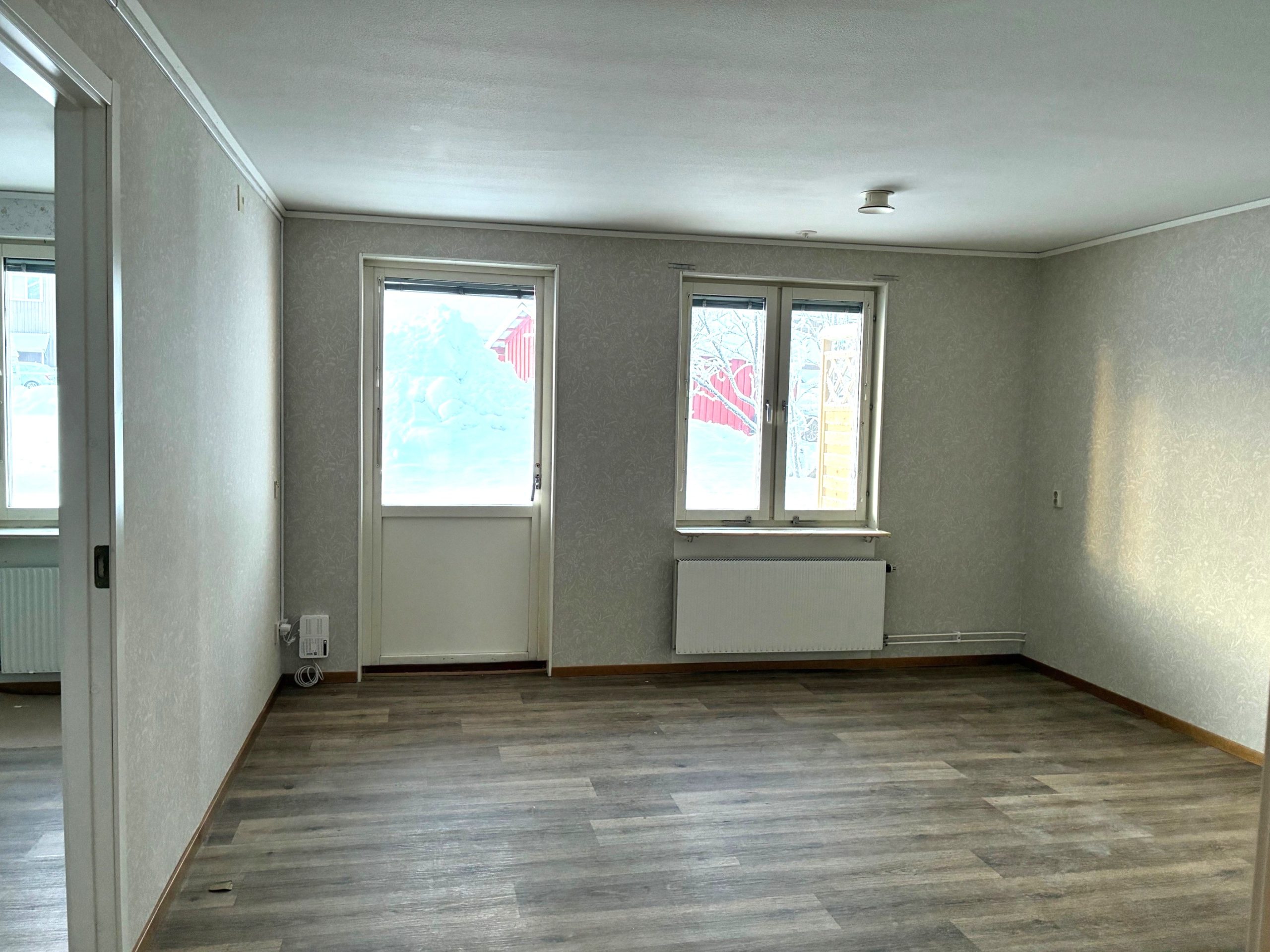 Vardagsrum med ljusa tapeter och träfärgat golv. På den bakre väggen finns ett fönster och en balkongdörr som släpper in ljus.