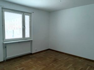 Vardagsrum med vita väggar, parkettgolv, samt ett fönster i bakgrunden.