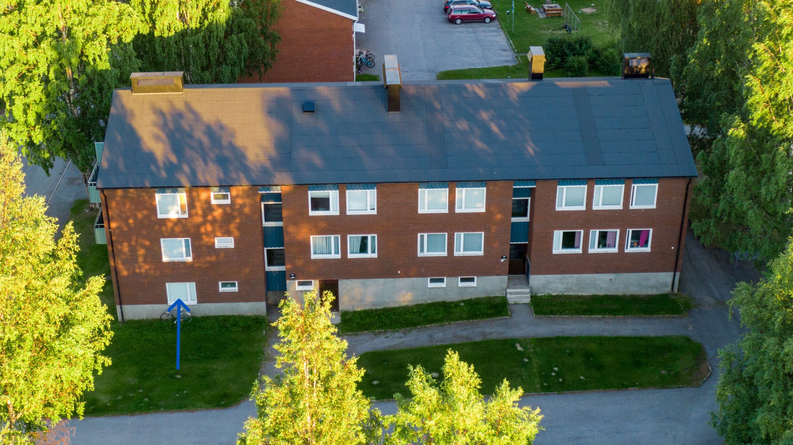 Ett brunt lägenhetshus med svart tak med fönster. En blå pil pekar mot en specifik lägenhet i fastigheten,